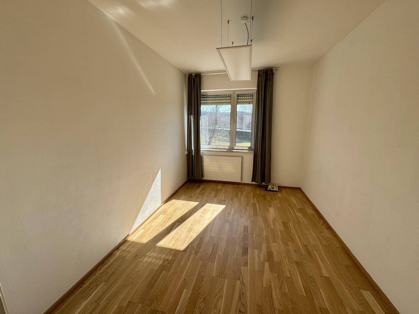 Helles Zimmer mit Parkettboden und Fenster, das viel natürliches Licht hereinlässt.