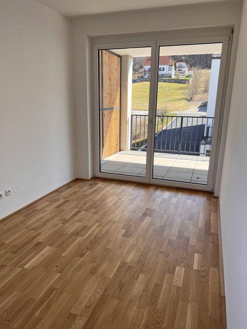 Helles Schlafzimmer mit Parkettboden und Zugang zu einem Balkon mit Sichtschutz.