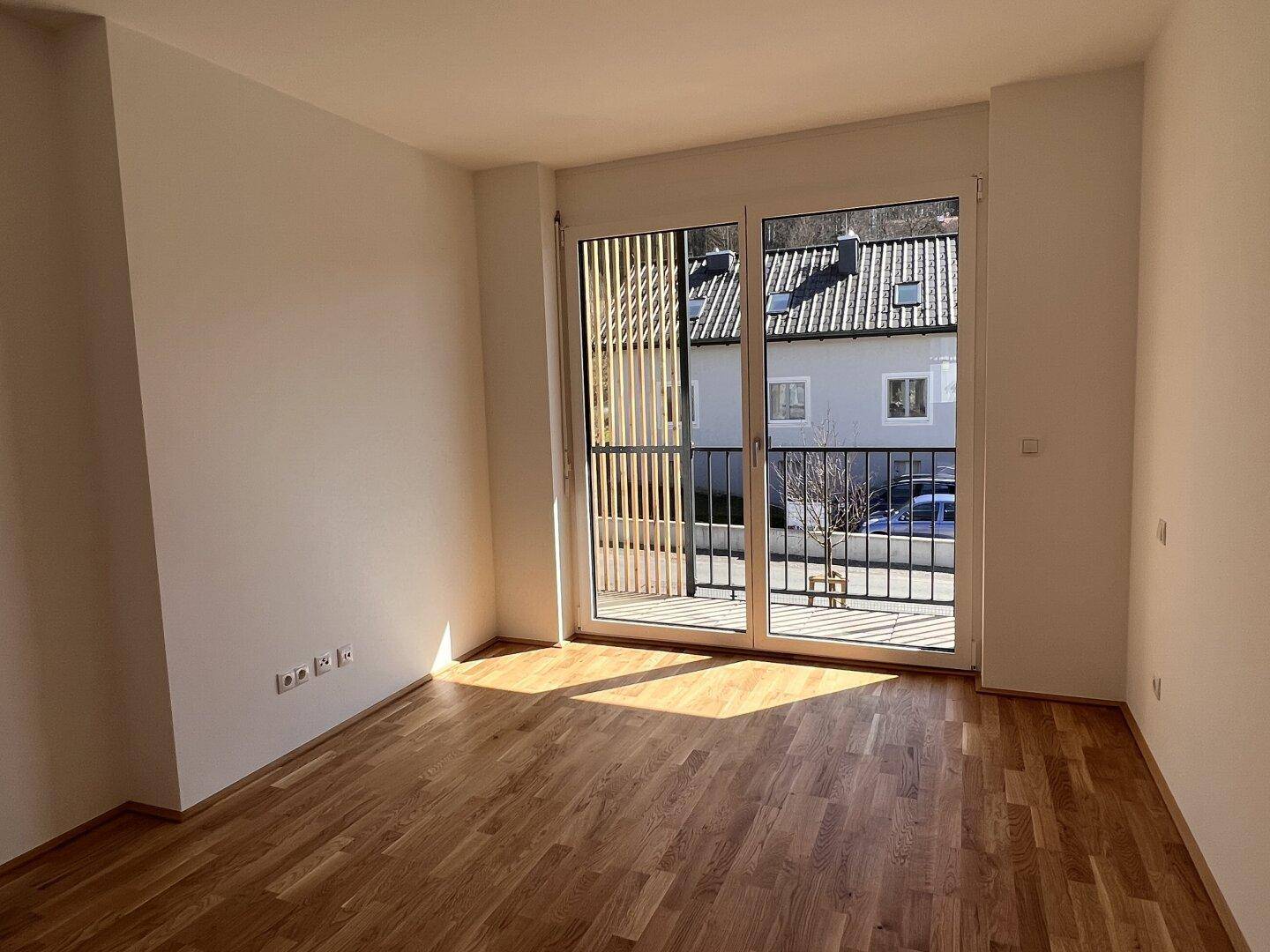 Lichtdurchfluteter Raum mit Holzboden und großen Fenstern, die Zugang zu einem Balkon bieten.