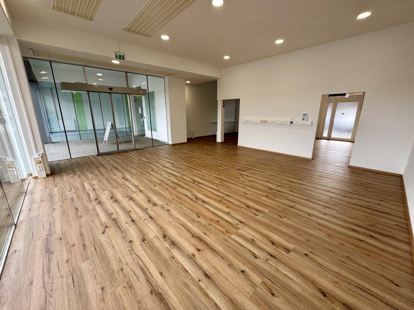 Heller Gewerberaum mit großen Glasfronten und neuem Laminatboden, ideal für ein Geschäft oder Büro.