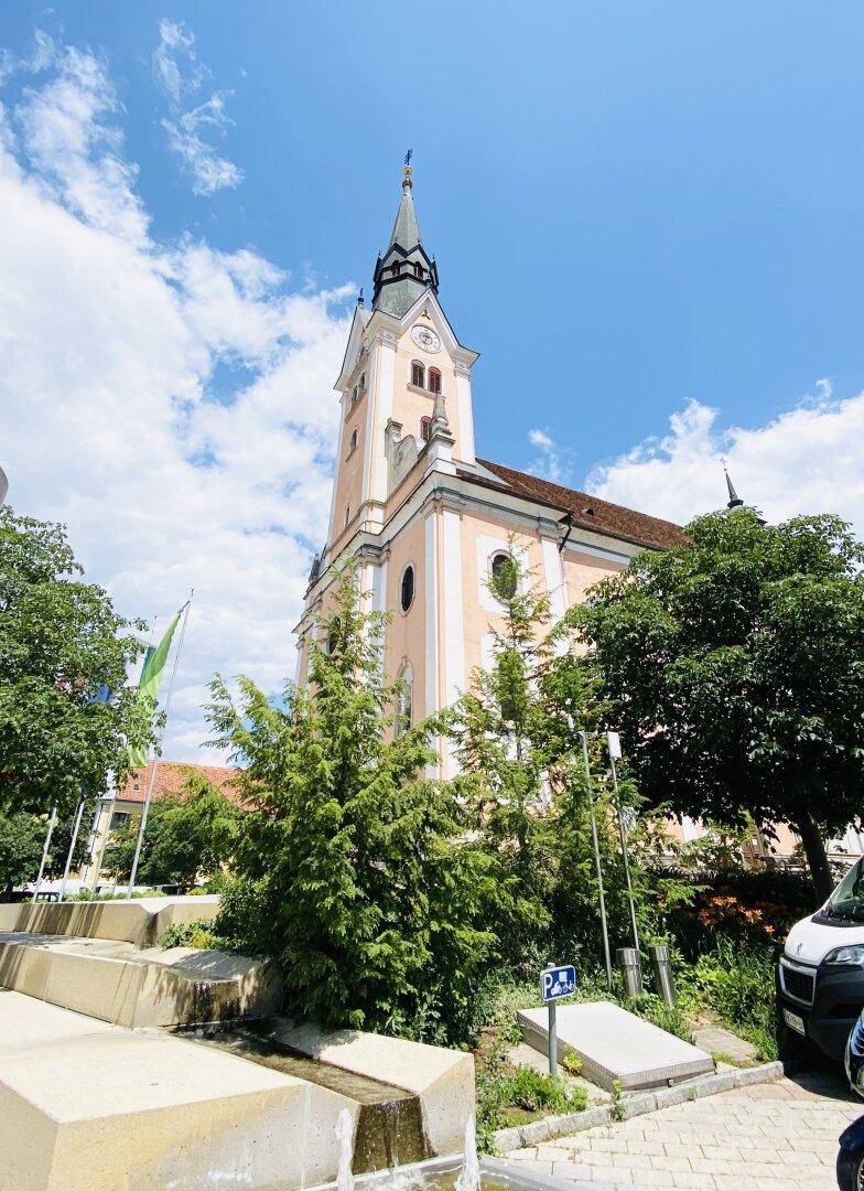 Historische Kirche mit hohem Turm und grünen Bäumen unter einem bewölkten Sommerhimmel.