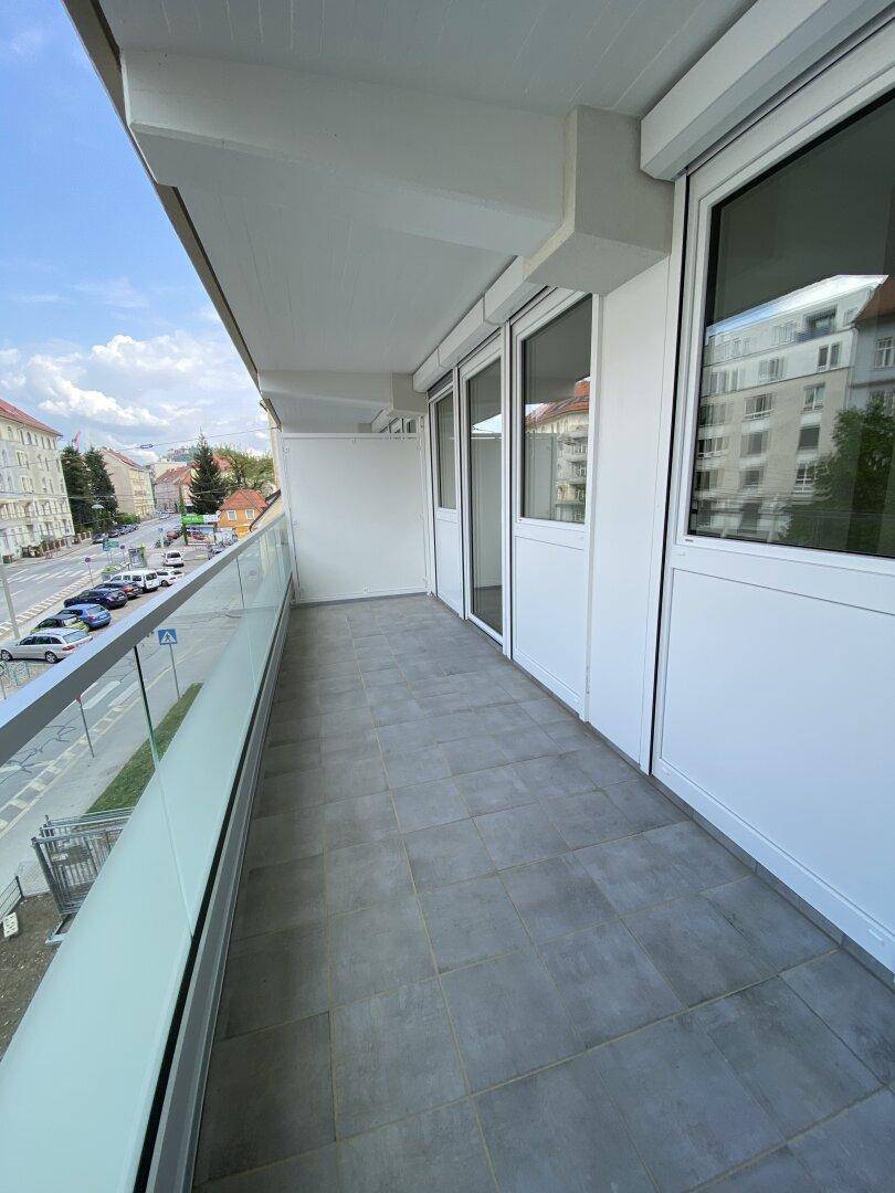Langer Balkon mit Fliesenboden und Glasbrüstung, mit Blick auf die Straße.
