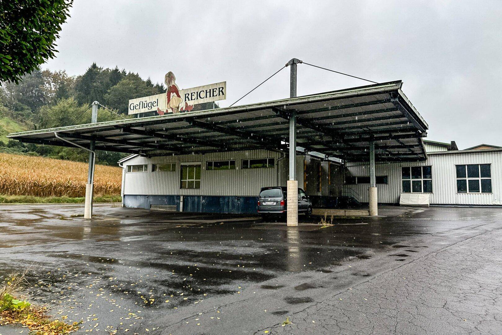 Außenansicht eines Gewerbegebäudes mit großem Vordach und Parkplatz, umgeben von Feldern bei Regenwetter.
