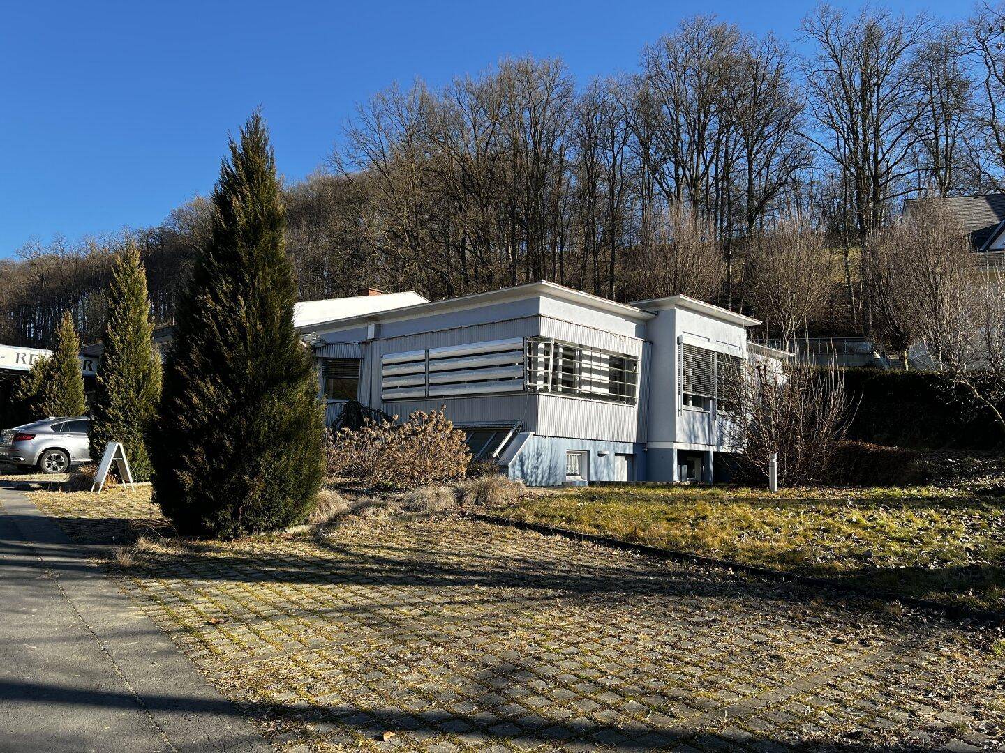 Modernes Gebäude mit grauer Fassade und horizontalen Lamellen, eingebettet in eine grüne Hanglage mit Bäumen.