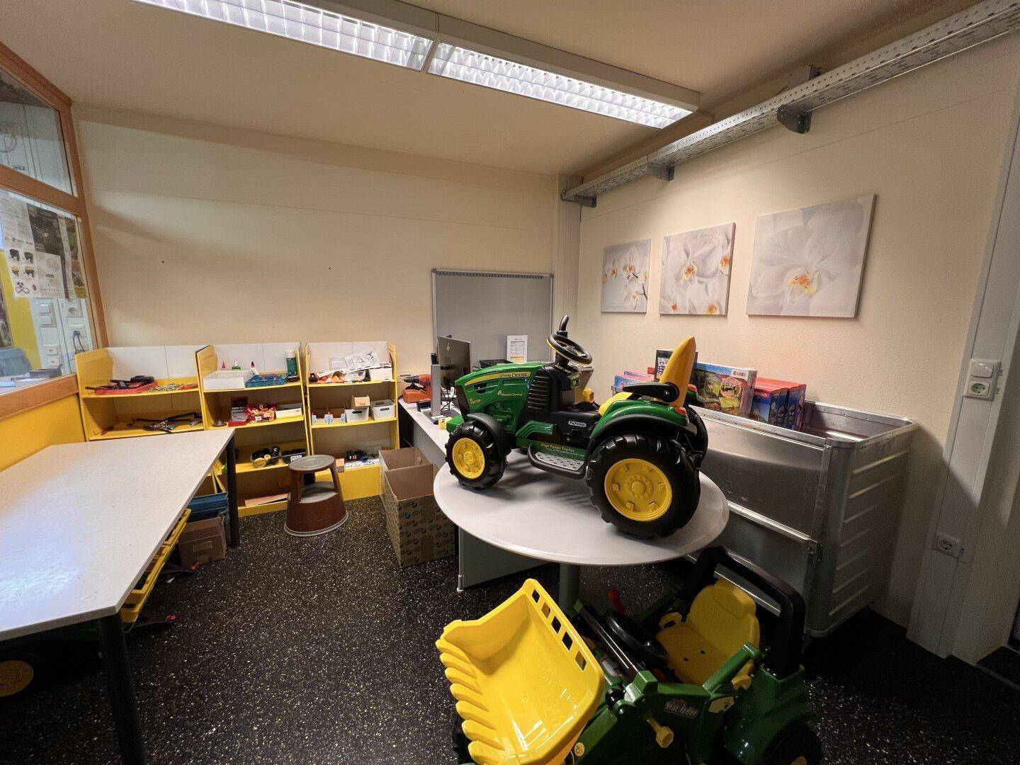 Raum mit Arbeitstisch, Regalen und einem großen Spielzeugtraktor, möglicherweise ein Lager oder Werkstatt.