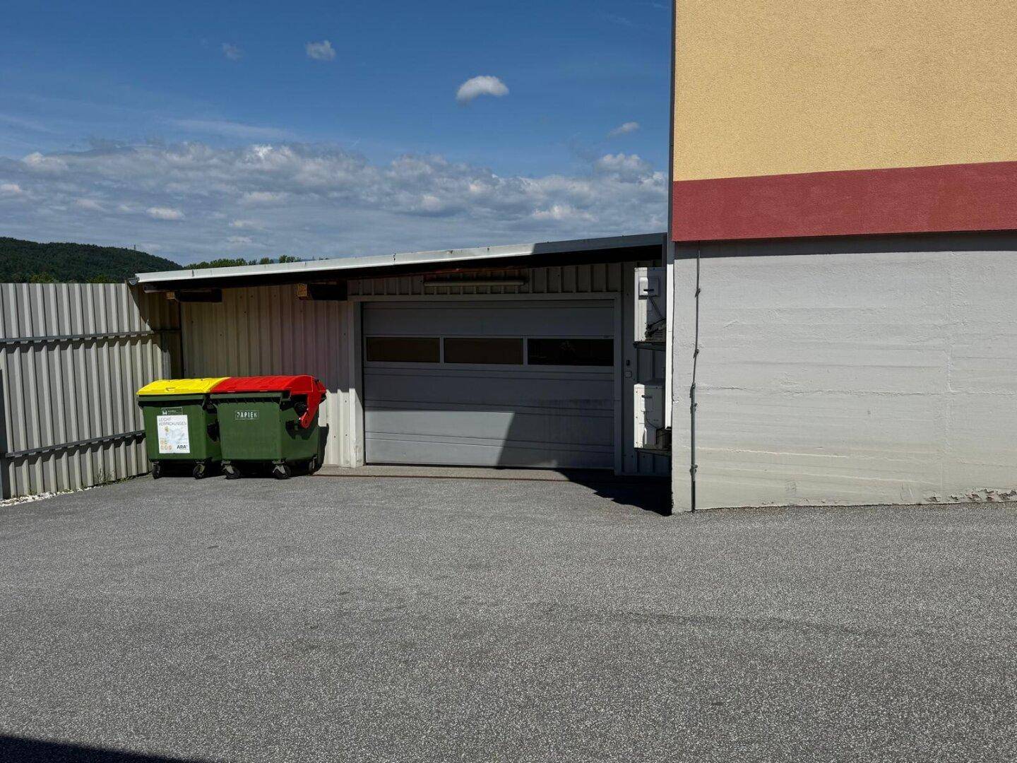 Überdachter Park- oder Lagerbereich mit Garagentor und Mülltonnen neben dem Gebäude.