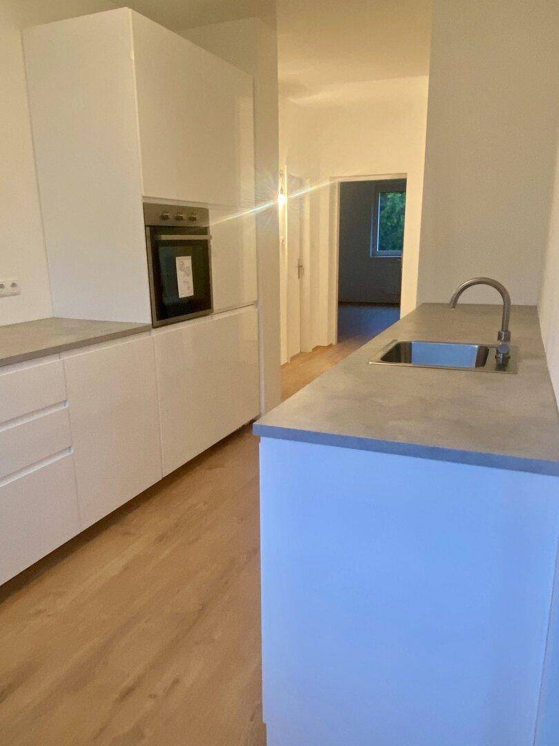 Helle, moderne Küche mit integriertem Backofen, Spülbecken und Zugang zu einem weiteren Raum.
