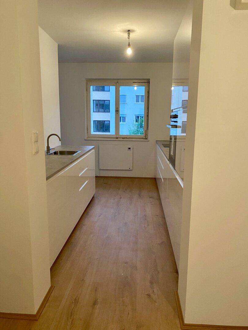 Moderne Küche mit weißen Schränken, grauen Arbeitsplatten und hellem Holzboden, mit Blick aus dem Fenster.
