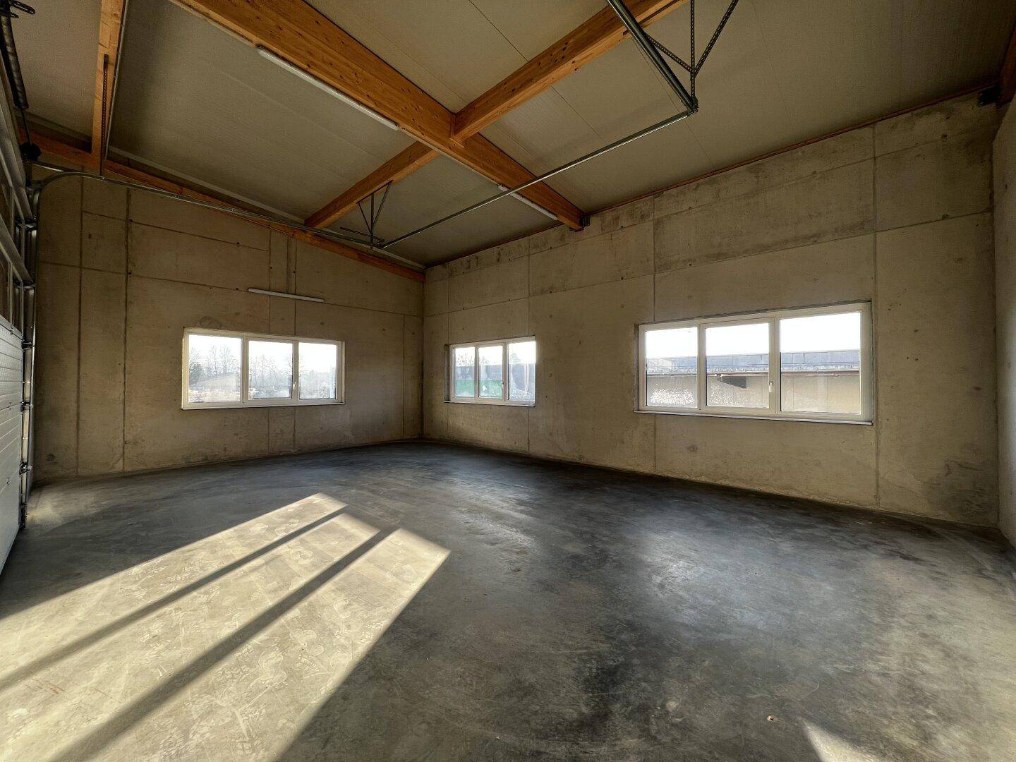 Großer, leerer Innenraum mit Betonwänden, Holzbalken und viel Tageslicht durch die Fenster.