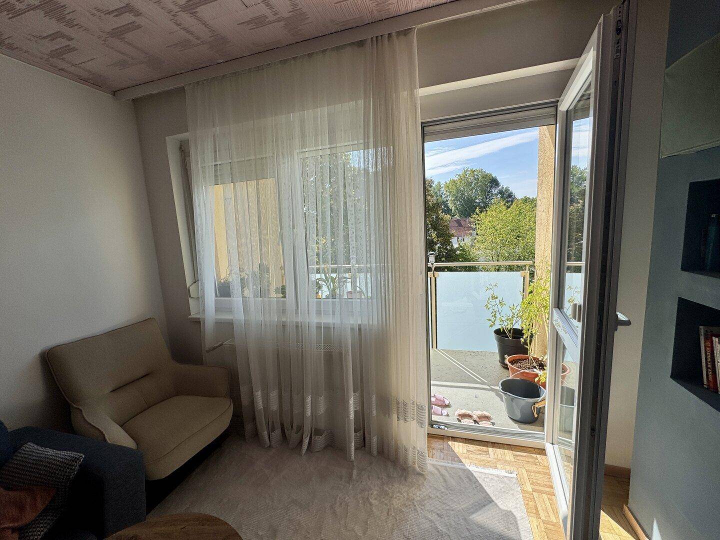 Heller Wohnbereich mit Zugang zu einem kleinen Balkon und Blick auf grüne Bäume.