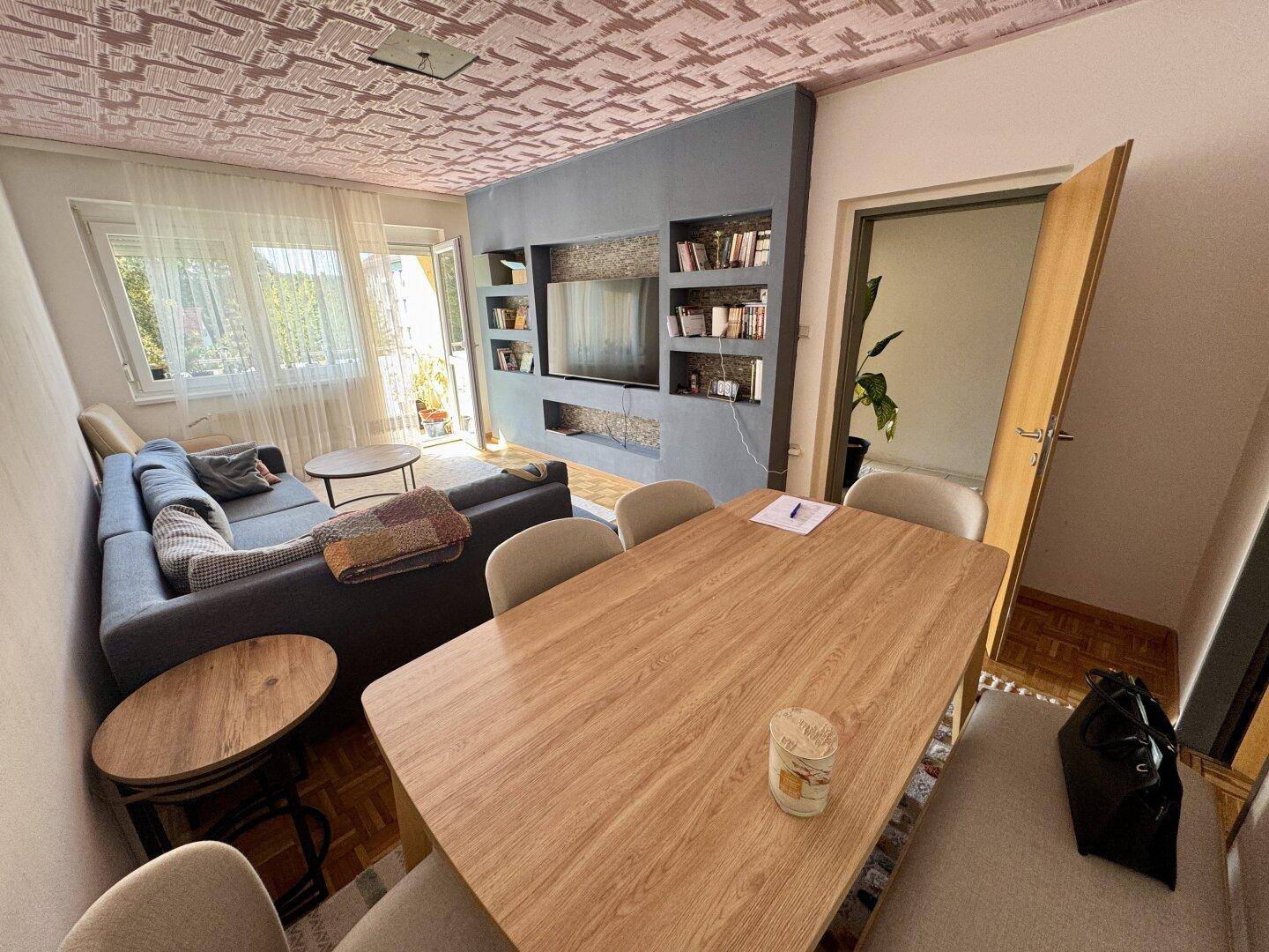 Gemütlicher Wohn- und Essbereich mit Holztisch, Sofa und Zugang zum Balkon, mit auffälliger Decke.