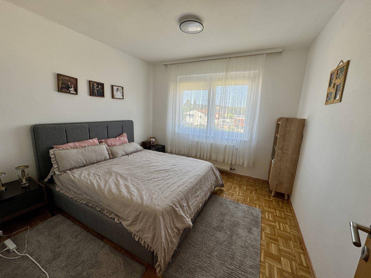 Schlafzimmer mit Doppelbett, Parkettboden und großem Fenster, das viel Tageslicht hereinlässt.