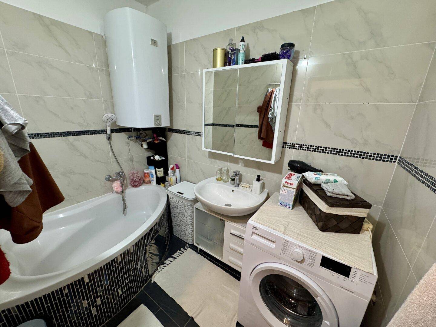 Modern gefliestes Badezimmer mit Badewanne, Waschbecken und Waschmaschine, gut beleuchtet.