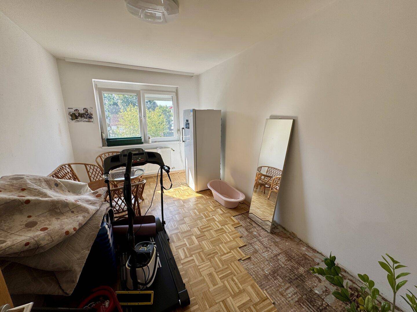 Schlafzimmer mit Parkettboden, Fenster und Fitnessgerät, teilweise renovierungsbedürftig.