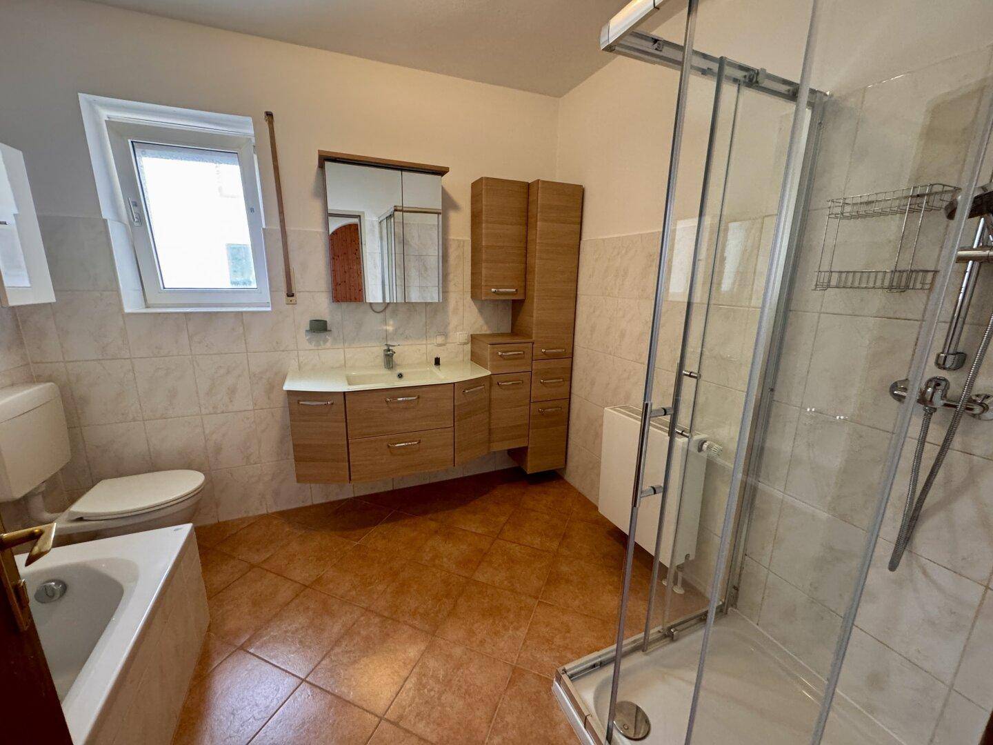 Badezimmer mit Badewanne, Duschkabine, Holzwaschtisch und Fenster, gefliest in Beige und Braun.