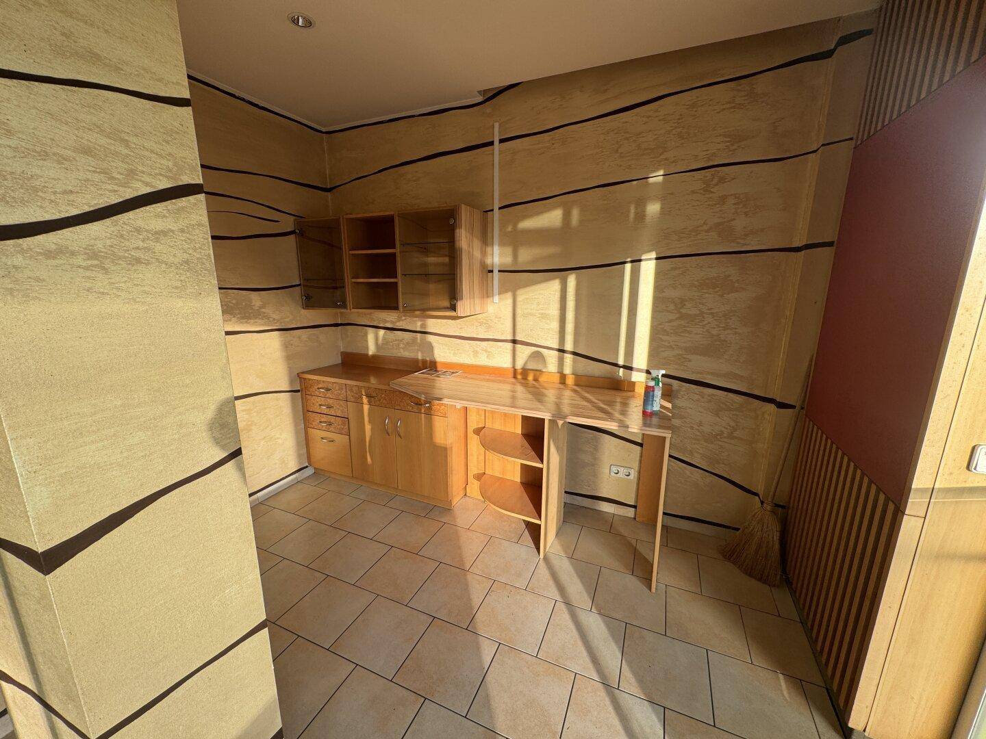 Ein kleiner Bürobereich mit Holztisch und Hängeschränken, beleuchtet durch natürliches Sonnenlicht.