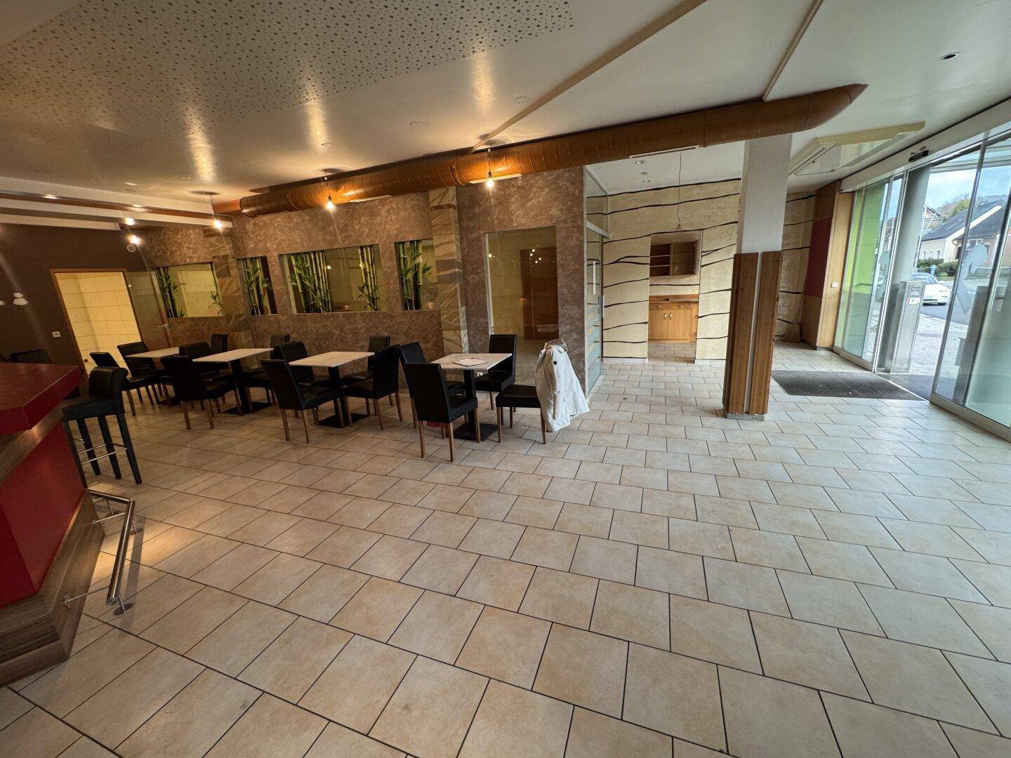 Ein heller Restaurantbereich mit Tischen und Stühlen, großen Fenstern und Zugang zur Straße.