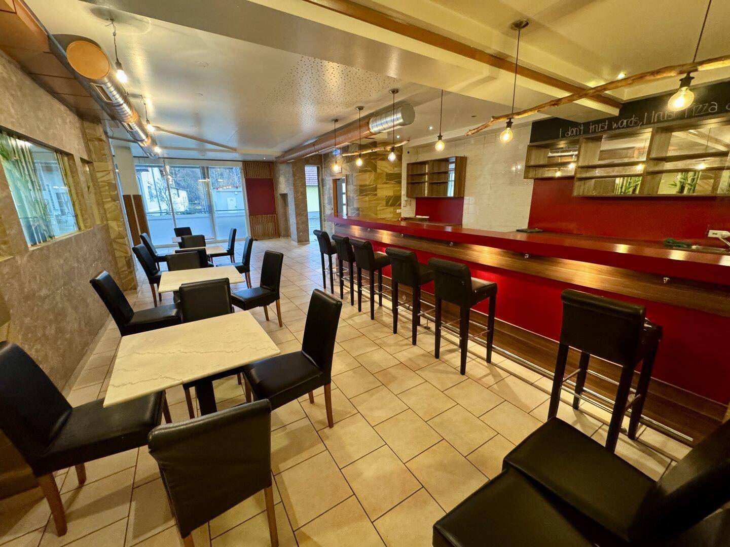 Ein geräumiger Restaurantbereich mit Tischen, Stühlen und einer langen Bar, beleuchtet durch moderne Hängelampen.
