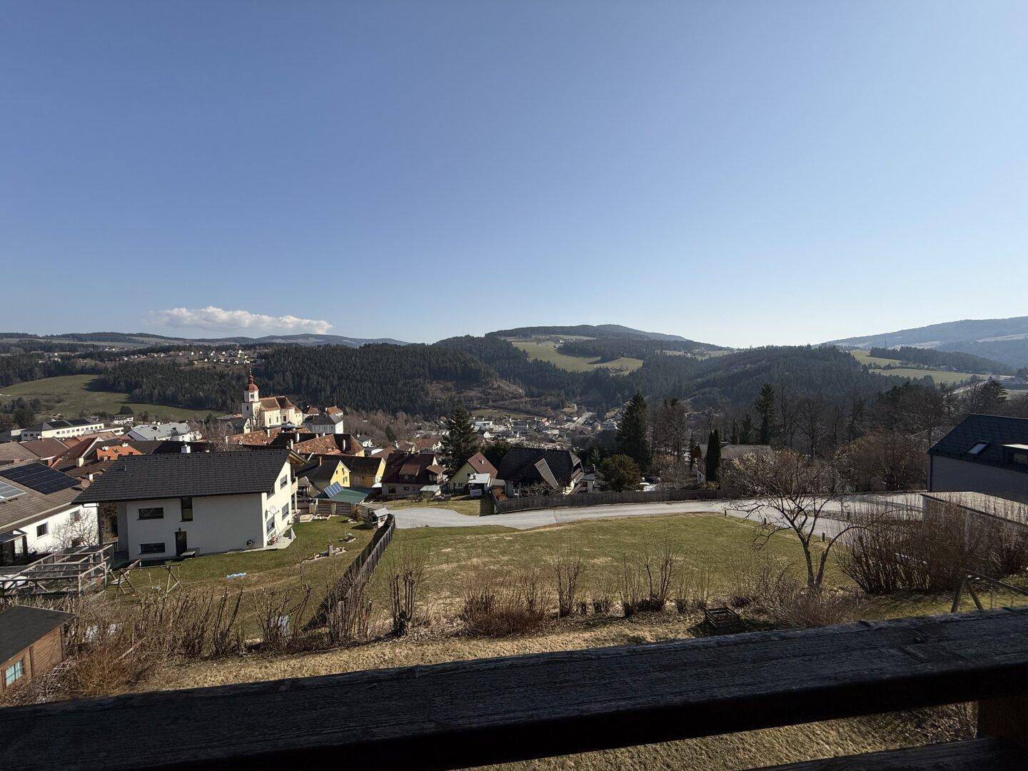 Panoramablick vom Balkon über das Tal, das Dorf und die umliegenden Berge unter blauem Himmel.