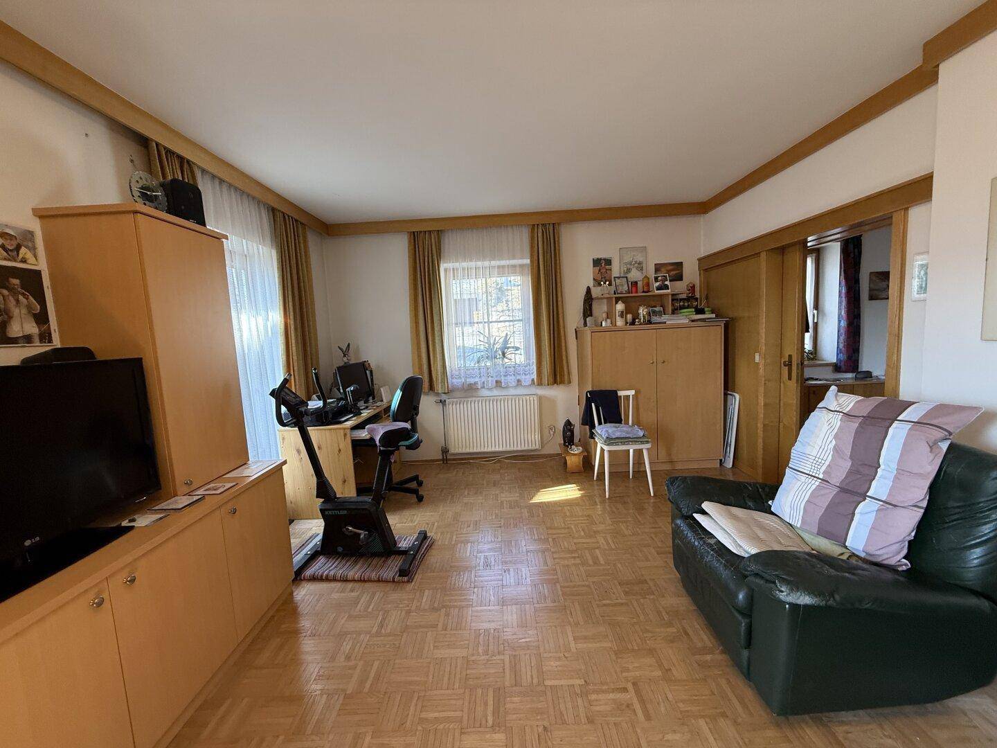 Helles Wohnzimmer mit Parkettboden, Fenster und Zugang zu einem weiteren Raum, ideal für Familien.