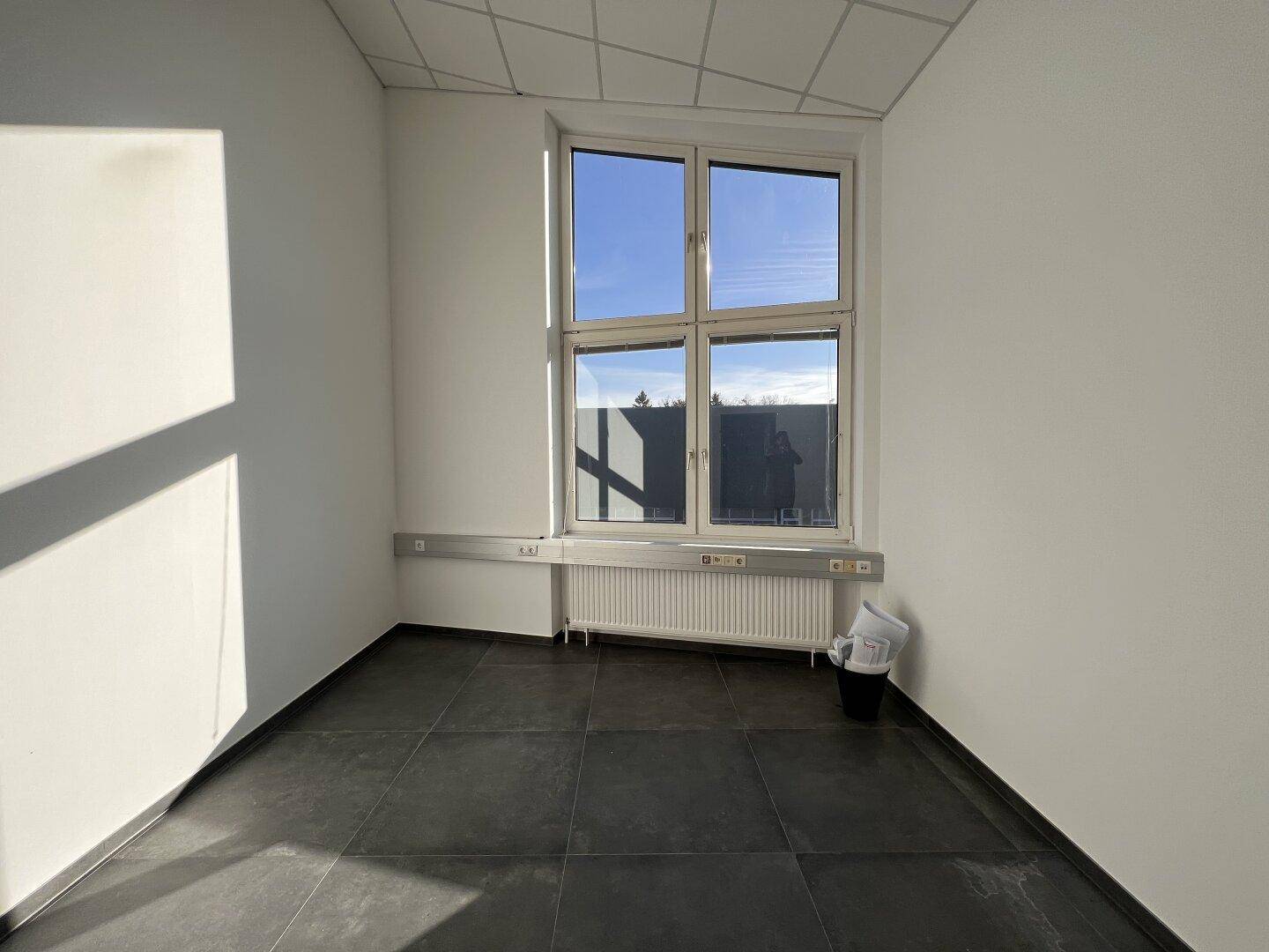 Heller, leerer Raum mit großen Fenstern, dunklen Fliesen und abgehängter Decke, durchflutet von Sonnenlicht.