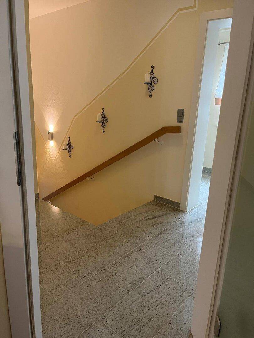 Flur mit gefliestem Boden, Holzgeländer und Wandleuchten, die die Treppe beleuchten.