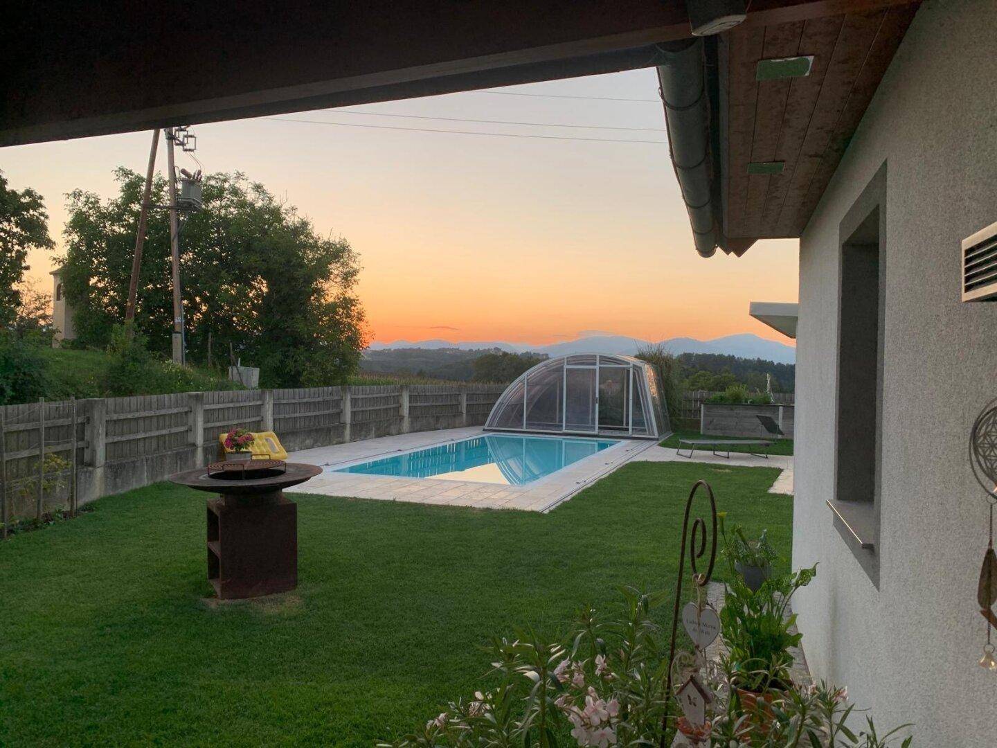 Abendliche Ansicht des Gartens mit beleuchtetem Pool, Rasen und Feuerstelle.