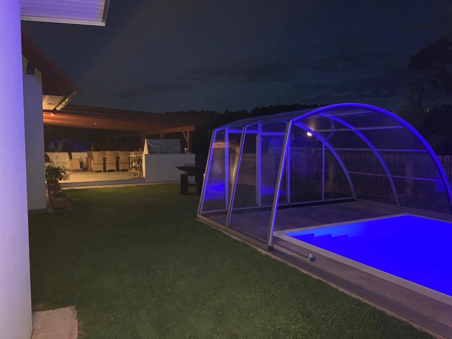 Nachtansicht des Gartens mit blau beleuchtetem Pool und überdachtem Terrassenbereich.
