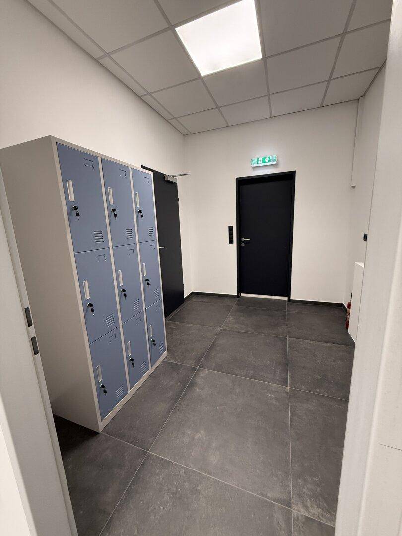 Heller Flur mit Schließfächern, dunklen Türen und Fliesenboden, der zu weiteren Räumen führt.