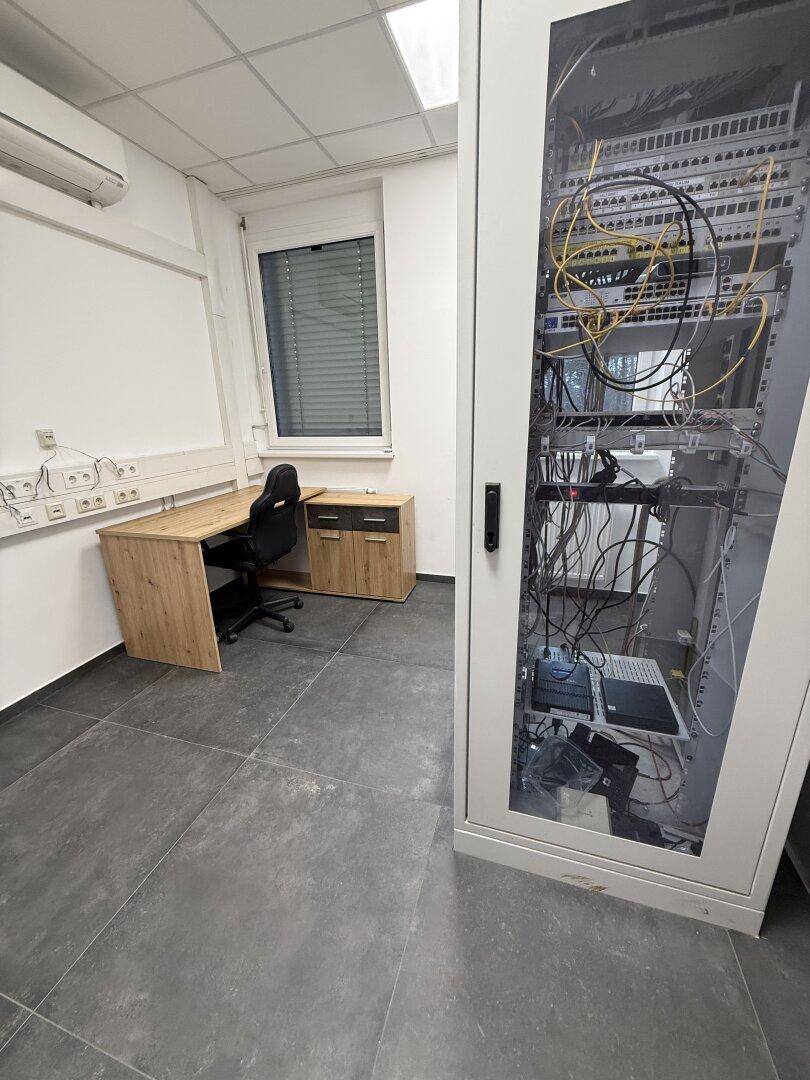 Büro- oder Serverraum mit Schreibtisch, Bürostuhl und einem großen Netzwerkschrank für IT-Infrastruktur.