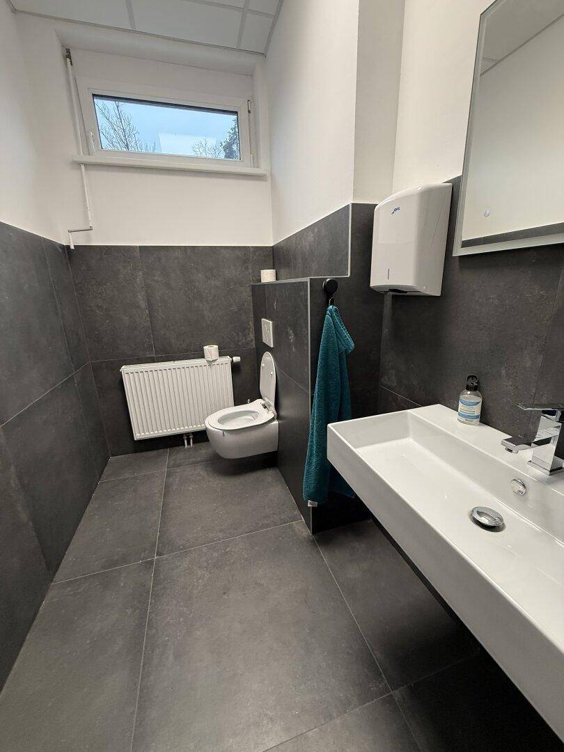 Modernes Badezimmer mit dunklen Fliesen, wandhängendem WC und großem Waschbecken mit Spiegel und Fenster.
