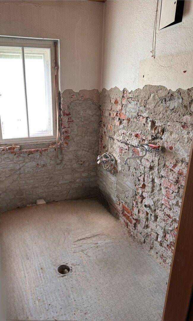 Badezimmer im Rohbauzustand mit freiliegenden Ziegelwänden und Fenster, bereit zur Renovierung.