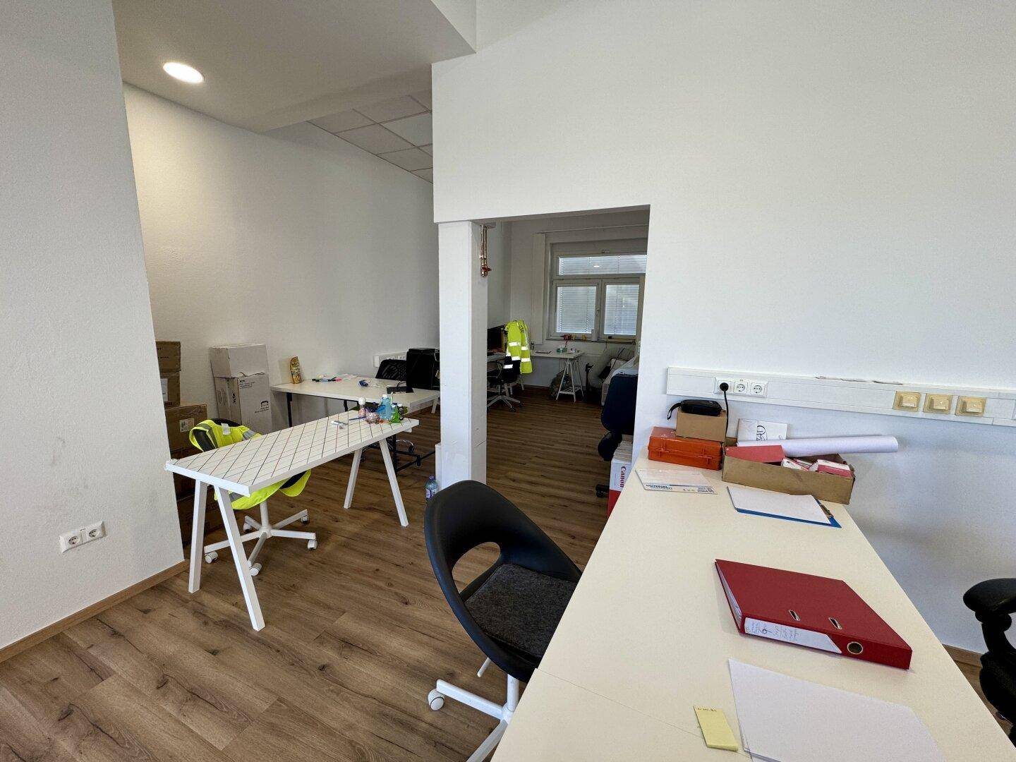 Heller Bürobereich mit schlichten Schreibtischen und Laminatboden, der zu einem weiteren Raum führt.