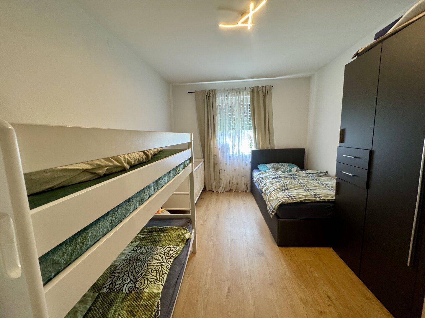 Kinderzimmer mit Etagenbett, Einzelbett und dunklem Kleiderschrank auf hellem Laminatboden.
