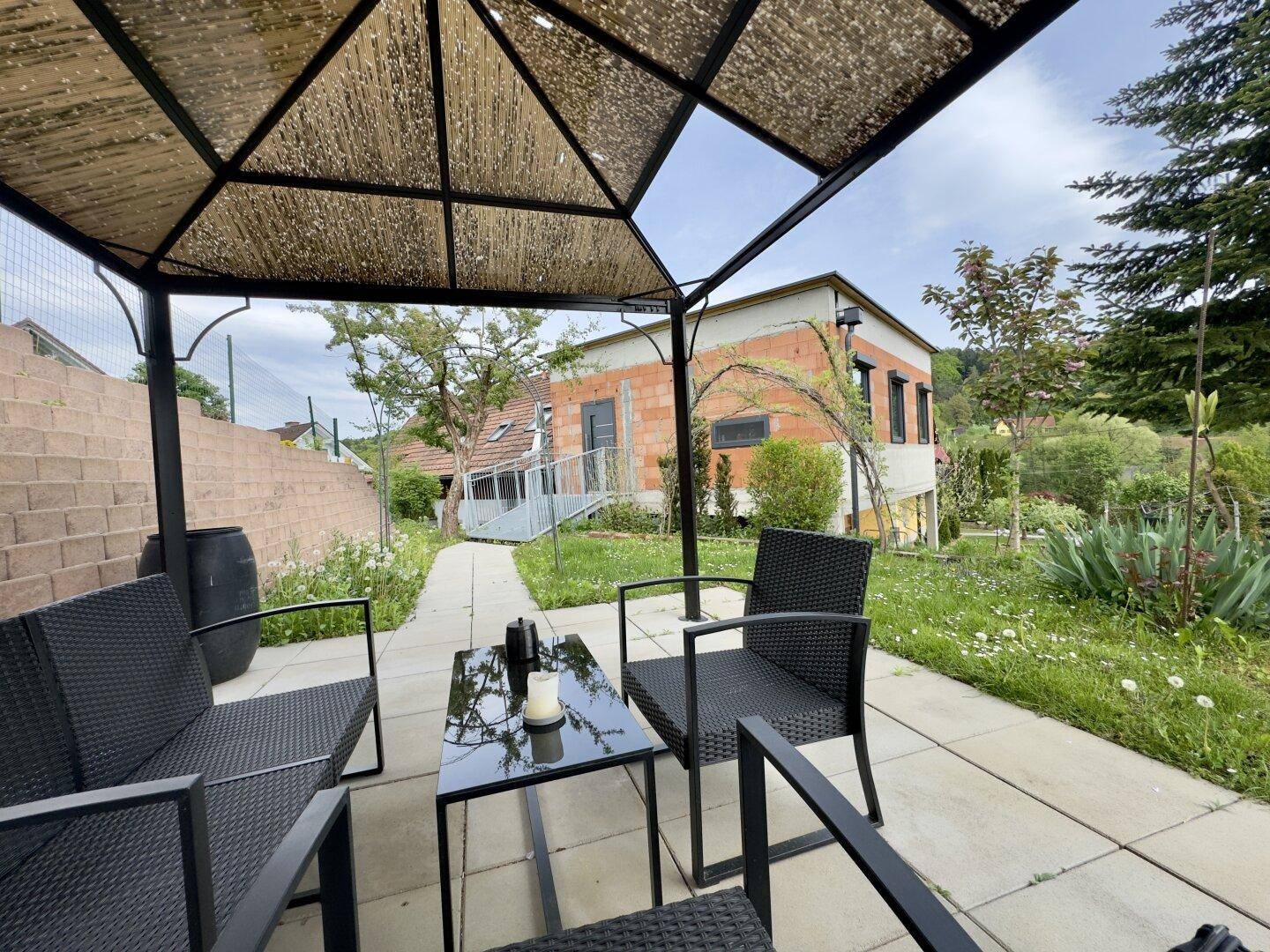 Gemütlicher Außenbereich unter einer Pergola mit Sitzgelegenheiten und Blick auf das Haus.