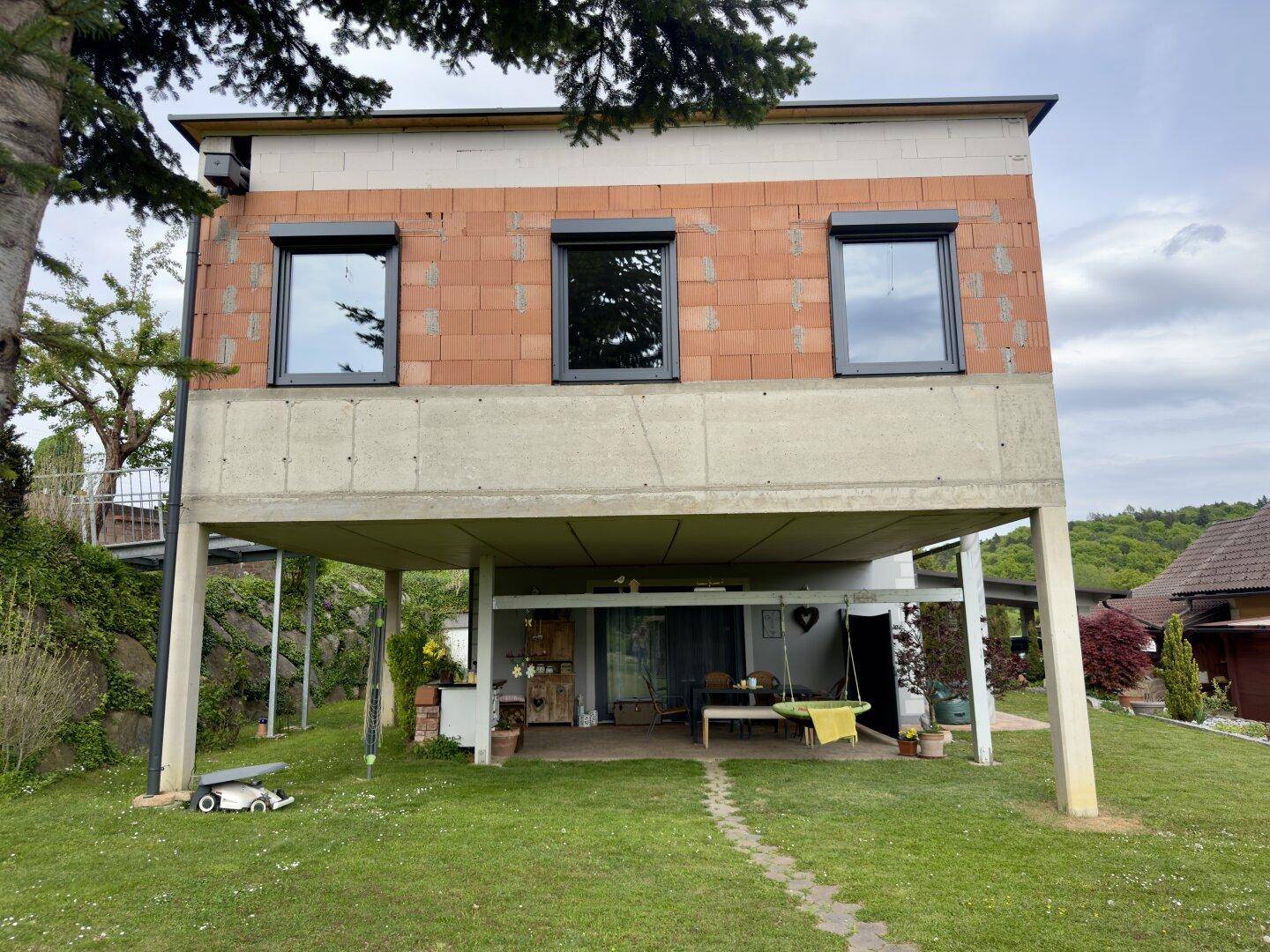 Seitenansicht des Hauses mit erhöhter Bauweise und überdachtem Außenbereich im Garten.