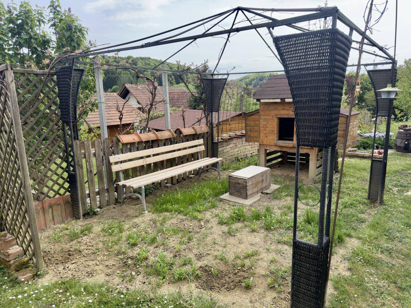 Außenbereich mit Holzbank, kleiner Hütte und Metallgestell, umgeben von Vegetation.