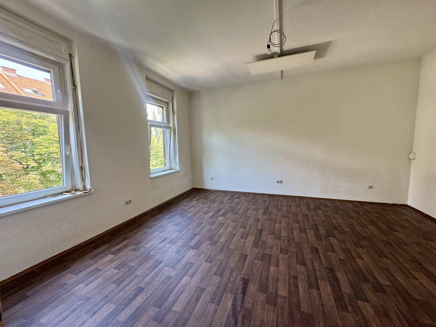 Heller Wohnraum mit dunklem Laminatboden und zwei großen Fenstern, die einen Blick ins Grüne bieten.
