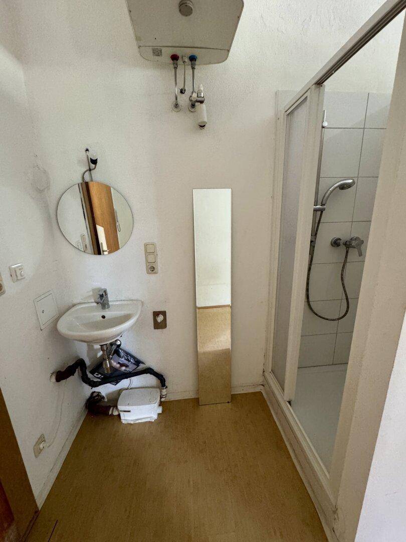 Kleines Badezimmer mit Duschkabine, Waschbecken, Spiegel und Warmwasserboiler an der Wand.