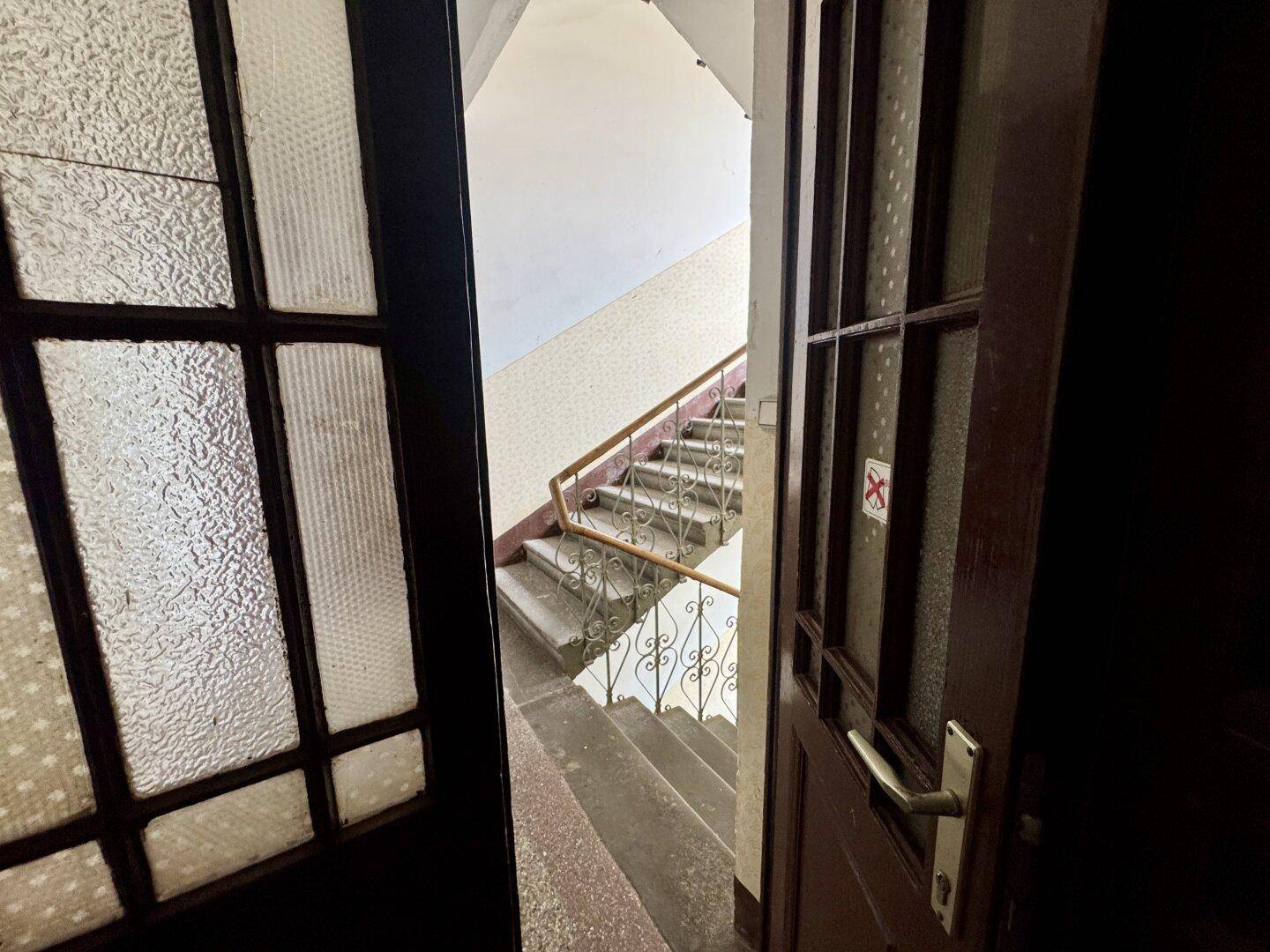 Blick durch eine offene Tür in ein Treppenhaus mit Steinstufen und einem verzierten Metallgeländer.