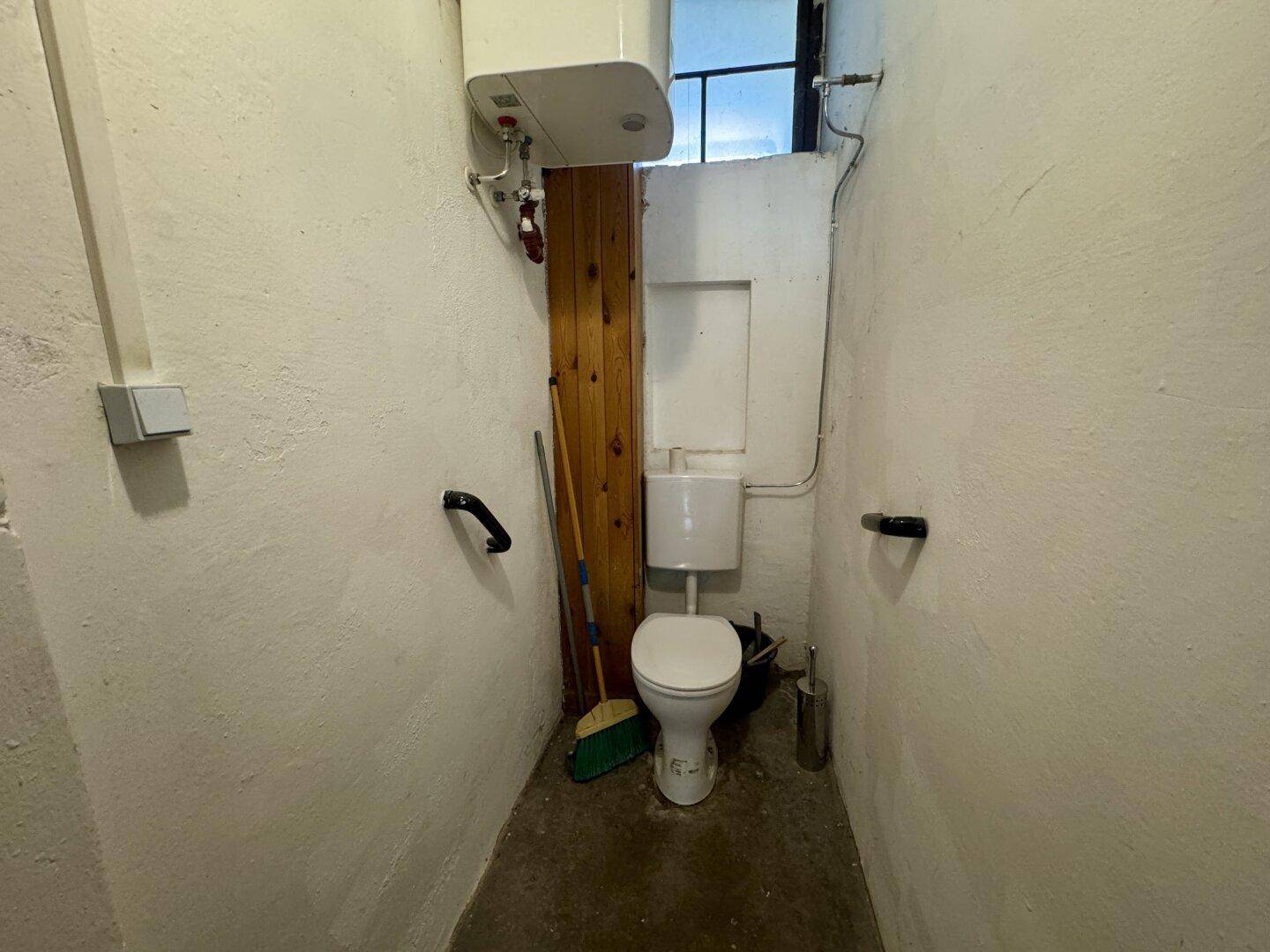 Kleines WC mit Toilette, Warmwasserboiler, Haltegriffen und einem kleinen Fenster für Belüftung.