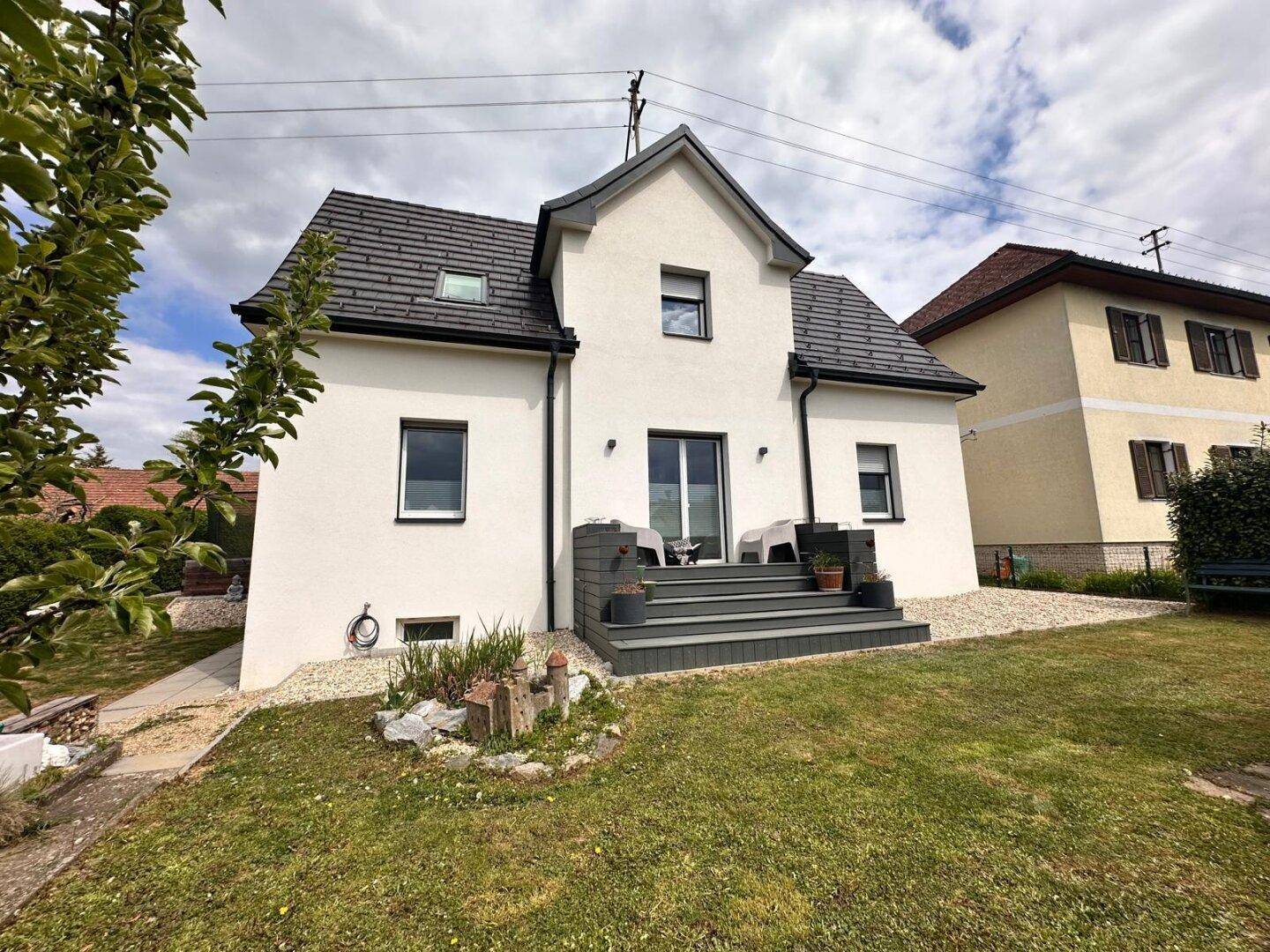 Modernes Einfamilienhaus mit weißer Fassade, dunklem Dach und kleiner Terrasse.