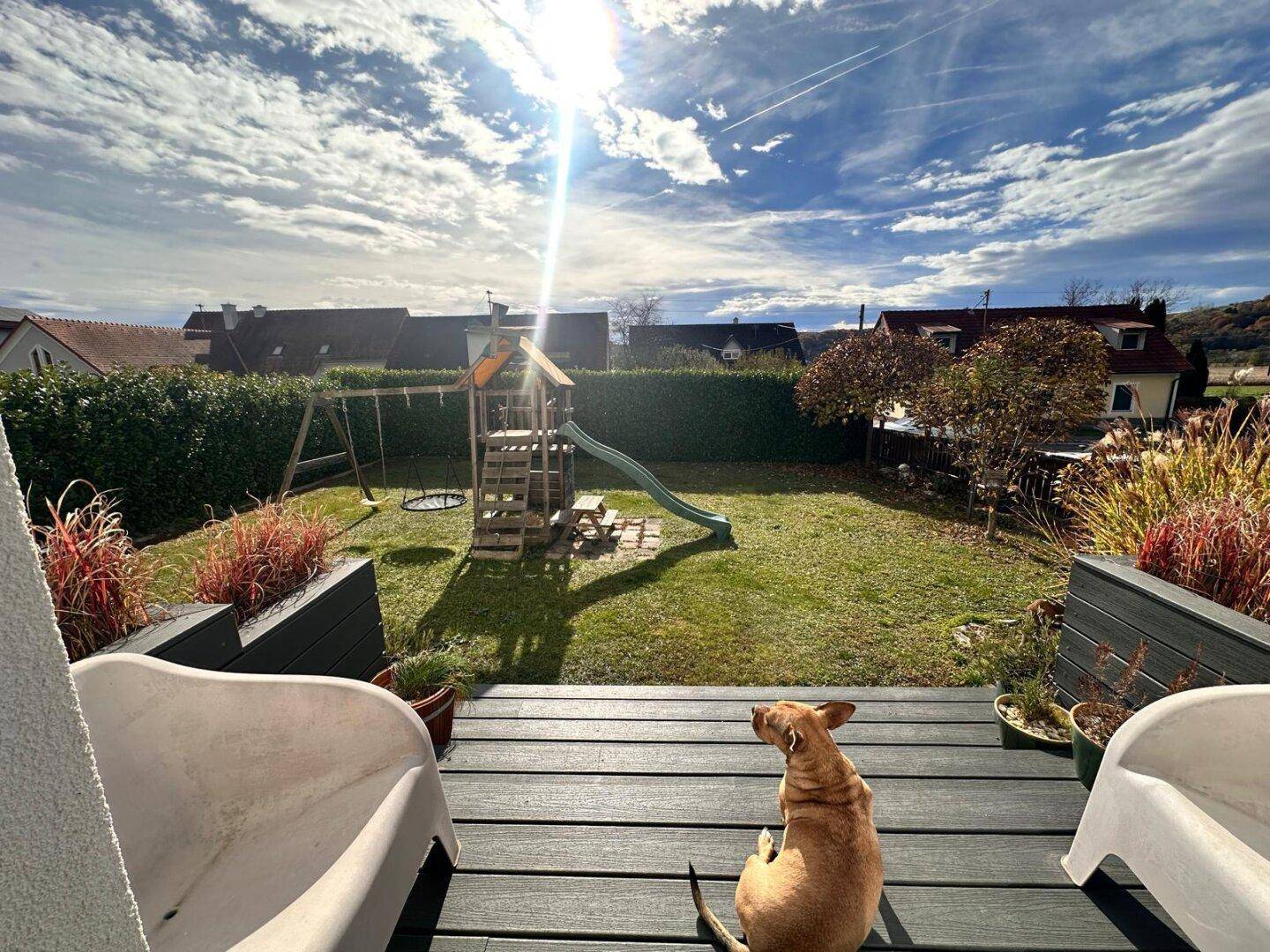 Sonniger Garten mit Holzterrasse, Spielplatz und Blick auf die Umgebung.