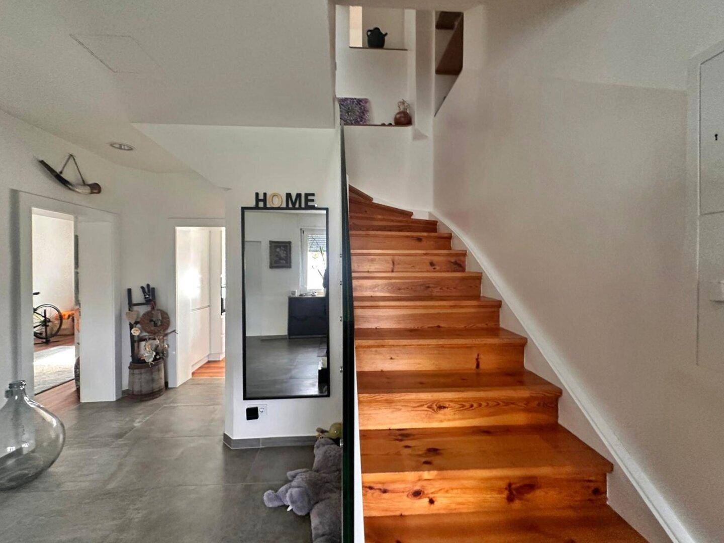 Heller Flur mit Holztreppe, modernem Spiegel und Fliesenboden.
