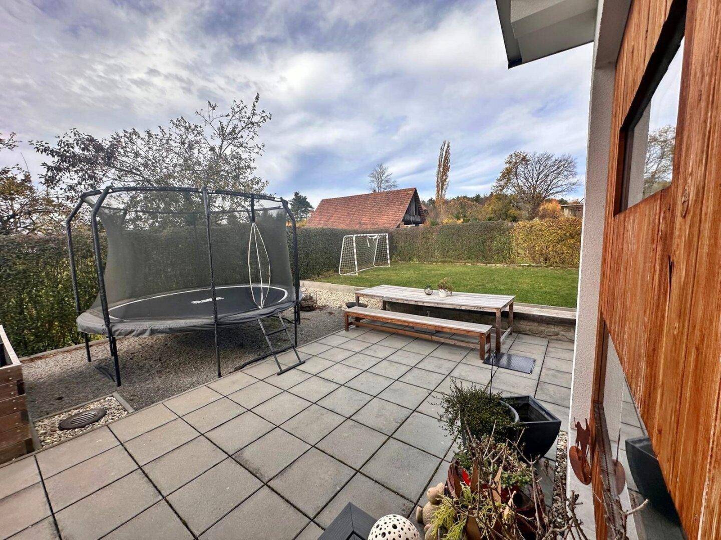 Große gepflasterte Terrasse mit Esstisch, Bänken und Trampolin im Garten.