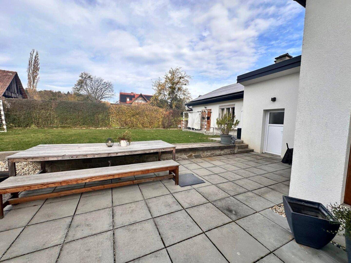 Geräumige gepflasterte Terrasse mit Holztisch und Bänken, angrenzend an den Garten.