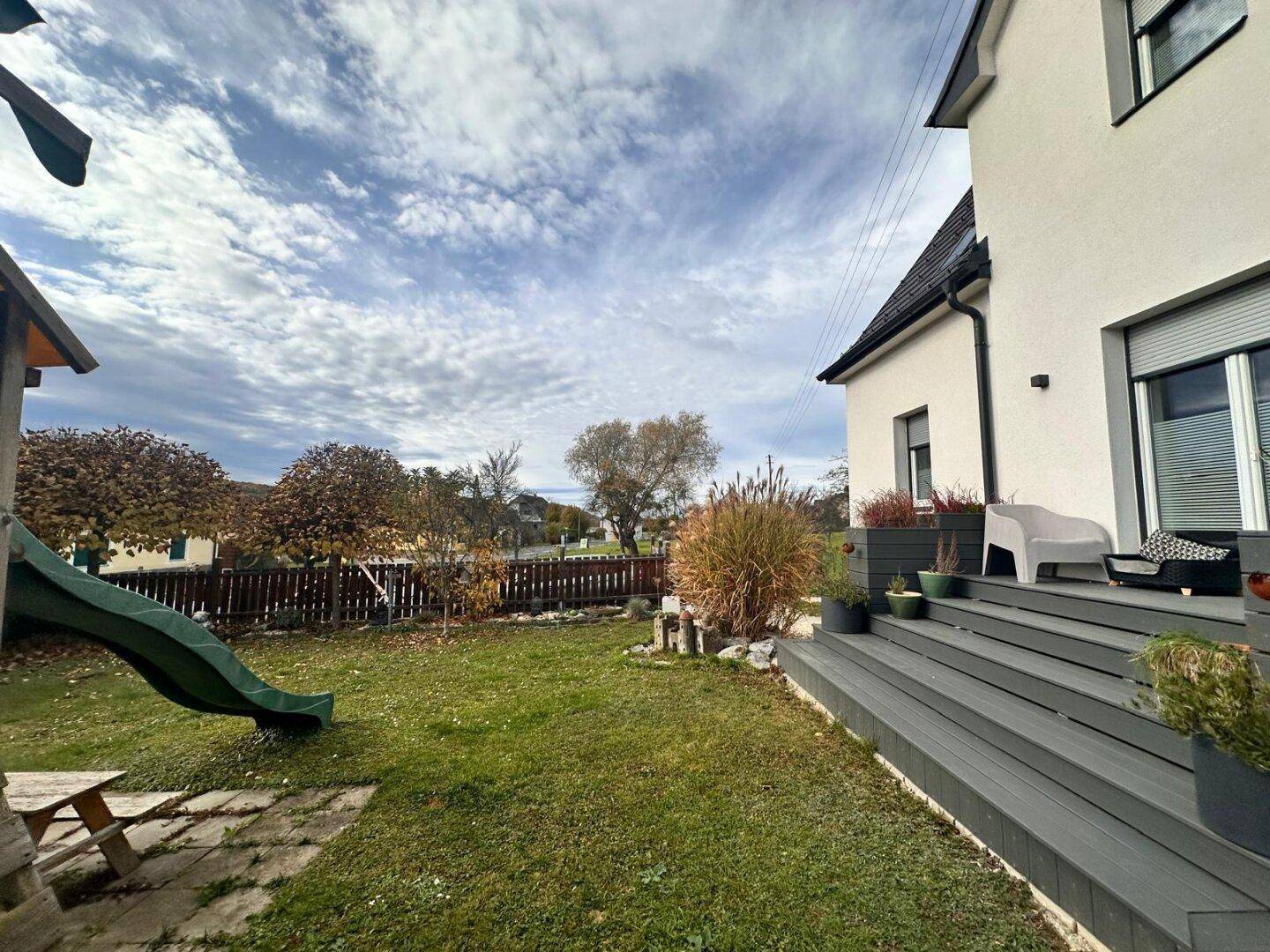 Weitläufiger Garten mit Spielplatz, grüner Hecke und angrenzender Holzterrasse.