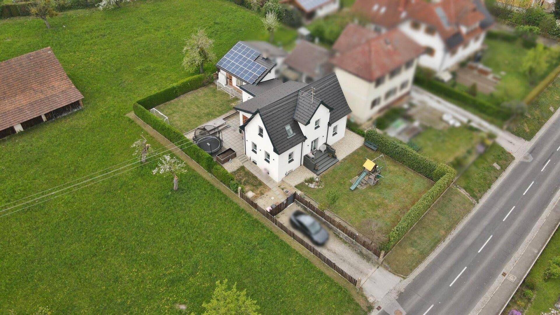 Luftaufnahme eines modernen Einfamilienhauses mit großem Garten, Spielplatz und Photovoltaikanlage auf dem Nebengebäude.