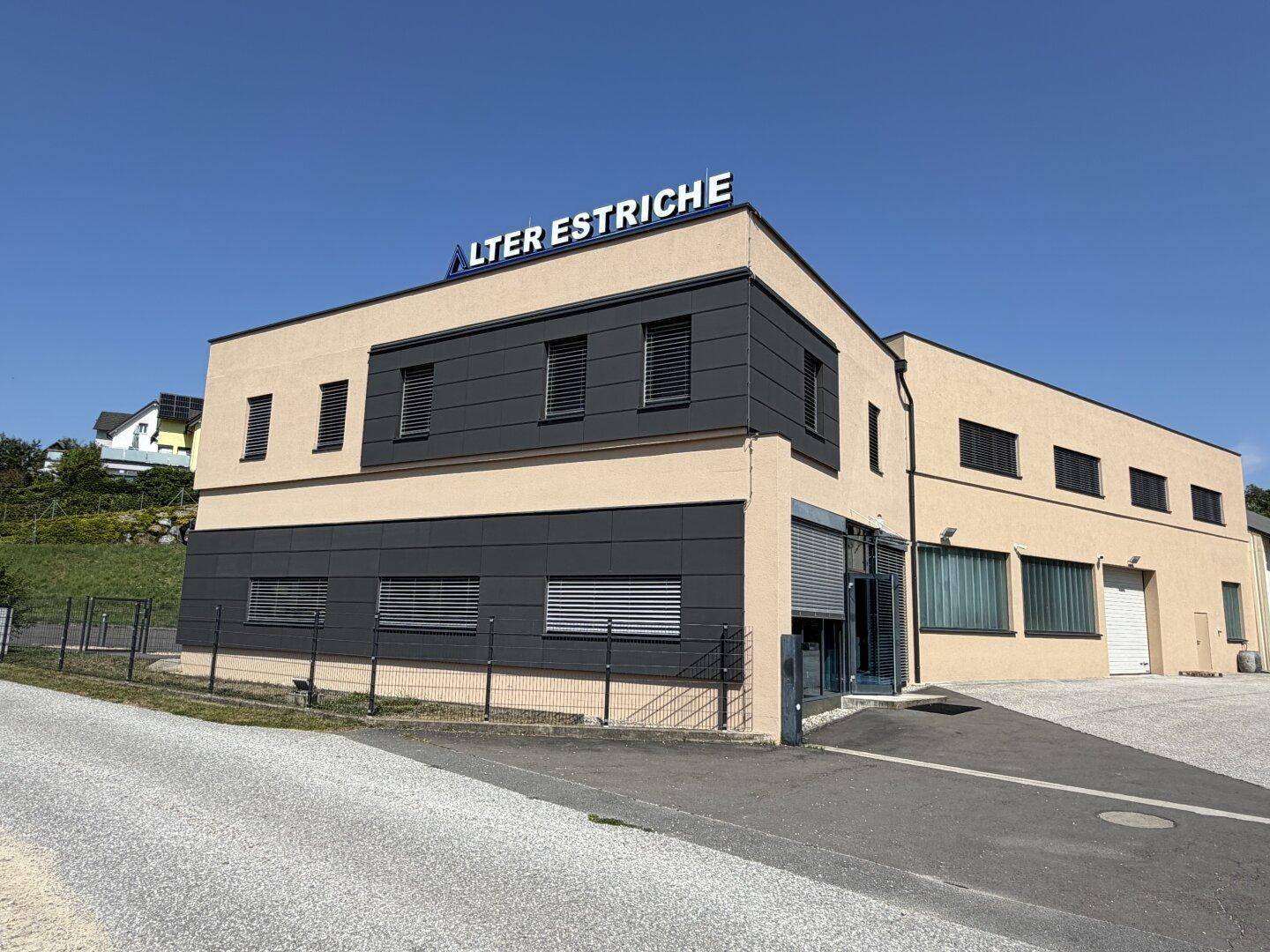 Modernes Geschäftsgebäude mit beiger und dunkelgrauer Fassade, Rollläden und einem Firmenschild unter blauem Himmel.