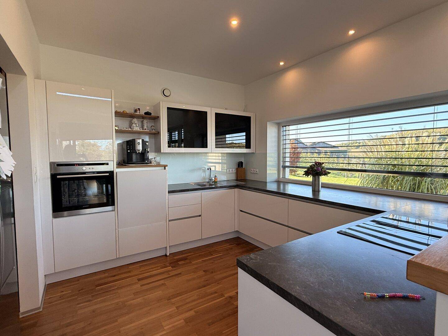 Moderne Einbauküche mit weißen Hochglanzfronten, dunkler Arbeitsplatte und Fensterblick.